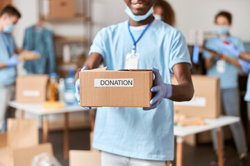 Wall Mural - Close up shot of cardboard box for Donation in hands of african american young male volunteer in blue uniform, protective mask and gloves. Team sorting, packing food stuff in the background