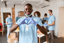 African American Young Male Volunteer In Blue Uniform, Protective Mask And Gloves Smiling At Camera And Showing Heart Love Sign. Team Sorting, Packing Food Stuff In The Background
