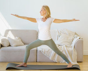 Calm and concentrated senior woman in home wear practicing Warrior Yoga Pose while exercising in morning at home, looking straight ahead, full length shot. Active and healthy lifestyle on retirement