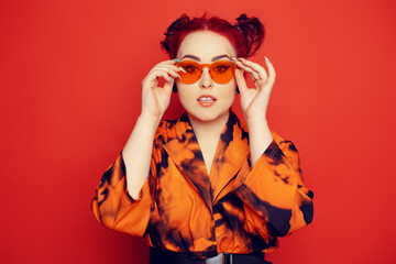 Cute girl on a red background. Red hair and tunnels in the ears, red dress and lips. Anime poses. A girl in orange sunglasses and two buns on her head looks at the camera.