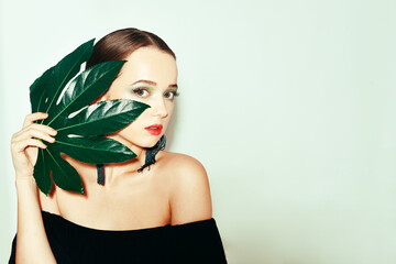 Wall Mural - Sexy beauty brunette model with green and blue make up and green leaves. Copy space. Close up, selective focus. Girl with tropical leaf covers a part of her face. Fashion, cosmetics, accessories.