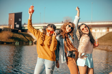 Wall Mural - Three girls near the river against the sky having fun. Fashion girls in sunglasses. Jump, rejoice, drink drinks, smile have fun, going crazy. Color dressed friends spend summer vibes, dance