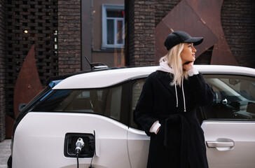 Young woman is standing near the electric car and looks at the smart phone. The rental car is charging at the charging station for electric vehicles. Car sharing.