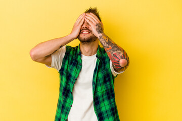 Young tattooed caucasian man isolated on yellow background laughs joyfully keeping hands on head. Happiness concept.