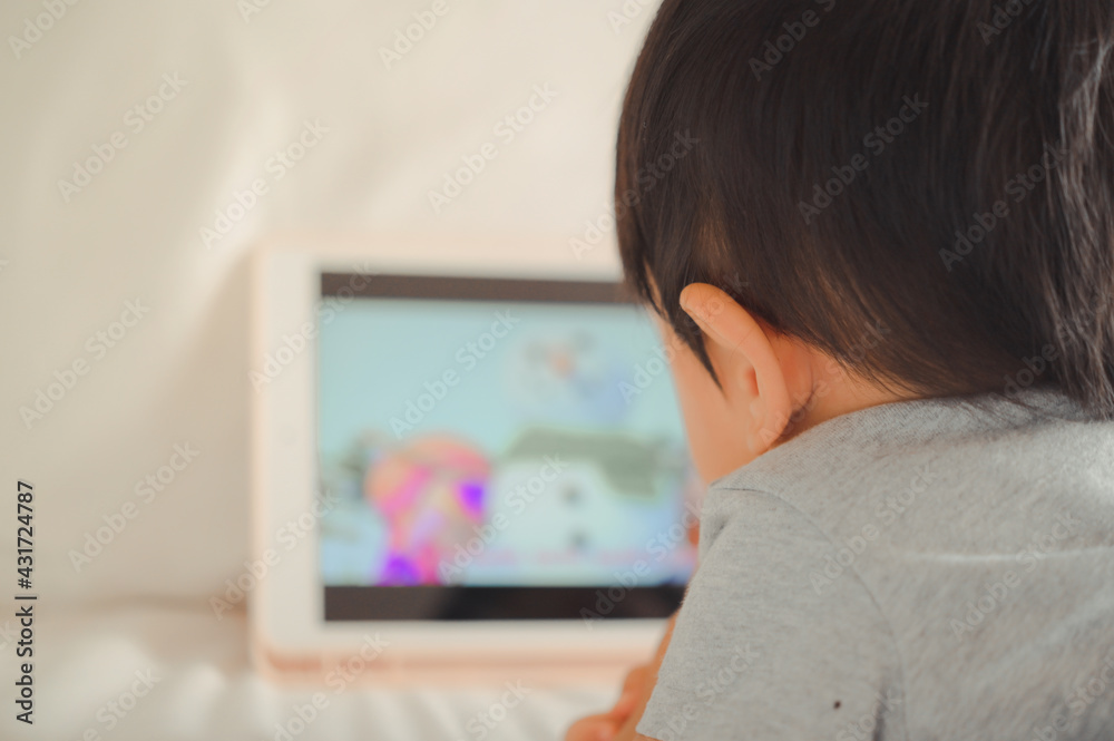 Little asian boy lying in bed and watch a story on touchscreen tablet ...