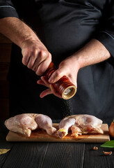 Chef or cook adds pepper to the chicken legs. Grill blank with raw chicken leg in the kitchen of a restaurant or cafe on a table with vegetables and spices