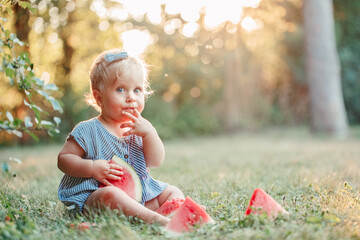 Summer picnic food. Cute Caucasian baby girl eating ripe red watermelon in park. Funny child kid sitting on ground with fresh fruit outdoor. Supplementary healthy finger food for toddler kids.