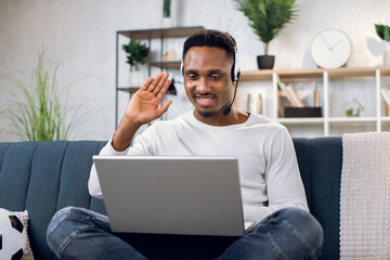 Wall Mural - Afro american male freelancer smiling and showing sign OK with fingers while having video conference in headset. Young man using wireless laptop for online meeting from home.