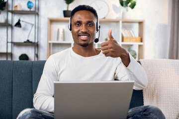 Wall Mural - Young black guy in headset sitting on couch with laptop, smiling and showing thumb up on camera. Male freelancer in casual clothes working remotely from home.