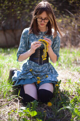 The girl sits on the grass and weaves a wreath of yellow dandelions