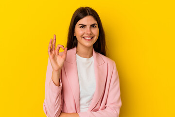 Young caucasian woman isolated on yellow background winks an eye and holds an okay gesture with hand.