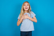Happy smiling beautiful Caucasian little girl wearing blue T-shirt over blue background has hands on chest near heart. Human emotions, real feelings and facial expression concept.