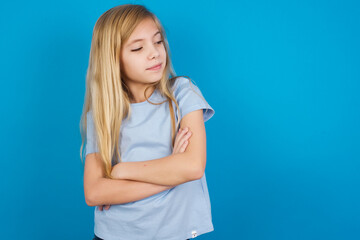 Wall Mural - Displeased beautiful Caucasian little girl wearing blue T-shirt over blue background with bad attitude, arms crossed looking sideways. Negative human emotion facial expression feelings.