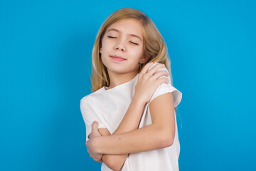 Wall Mural - beautiful Caucasian little girl wearing white T-shirt over blue background. Hugging oneself happy and positive, smiling confident. Self love and self care.