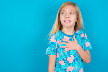 Joyful Caucasian kid girl wearing hawaiian T-shirt against blue wall expresses positive emotions recalls something funny keeps hand on chest and giggles happily.