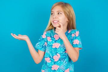 Wall Mural - Positive Caucasian kid girl wearing hawaiian T-shirt against blue wall advert promo touch finger teeth