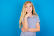 © Roquillo - Optimistic Caucasian kid girl wearing striped shirt ​against blue wall keeps hands partly crossed and hand under chin, looks at camera with pleasure. Happy emotions concept.