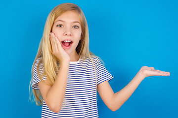 Wall Mural - Crazy Caucasian kid girl wearing striped shirt ​against blue wall advising discount prices hold open palm new product