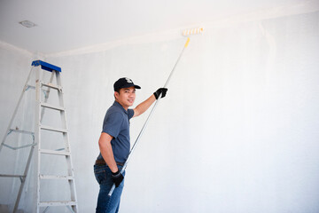 Wall Mural - The back view of an Asian painter paints the walls with a paint roller and a separate bucket on a large empty space with a wooden staircase Young worker painting wall in room