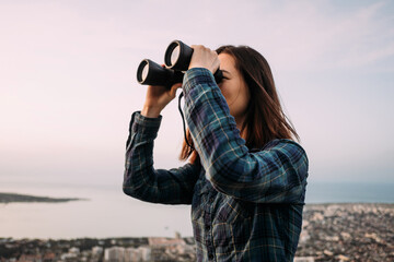 Poster - Portrait of a young female tourist looking through binoculars at sunset.