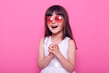 Charming female kid with dark hair wearing stylish heart shape glasses and white dress, looking directly at camera, keeps hands together, having amazing emotions, smiling happily.