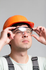 Wall Mural - protective glasses as element of builder's uniform in hands of young man, construction worker in helmet and overalls on grey background