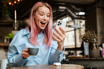 Wall Mural - Young excited woman using mobile phone while drinking coffee in cafe