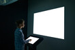 Mock up, futuristic, template and education concept. Woman using electronic kiosk and looking at white blank large interactive wall display in dark room of modern technology museum - mockup image