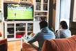 © Josu Acosta/Westend61 - Young male friends playing soccer game on television in living room at home