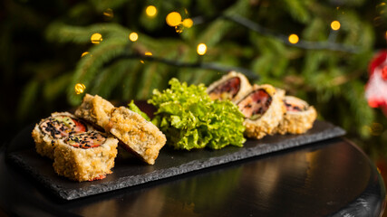 Wall Mural - Custom sushi roll in tempura with nori, fresh salmon, tuna, avocado, masago caviar, drizzled with pineapple sauce with salad pouring as decoration on a black plate on a wooden table for Christmas.