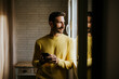 © Miguel Frias/Westend61 - Contemplating man looking through window while holding mobile phone at home