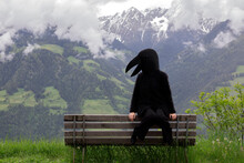 Female In Crow Costume Sitting On Bench Against Mountain Range