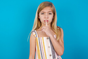 Wall Mural - beautiful Caucasian little girl wearing stripped dress over blue background makes hush gesture, asks be quiet. Don't tell my secret or not speak too loud, please!