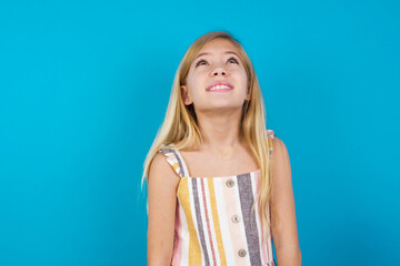 Wall Mural - Portrait of mysterious beautiful Caucasian little girl wearing stripped dress over blue background looking up with enigmatic smile. Advertisement concept.