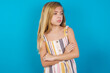 Displeased beautiful Caucasian little girl wearing stripped dress over blue background with bad attitude, arms crossed looking sideways. Negative human emotion facial expression feelings.