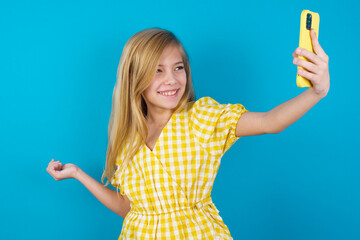 Wall Mural - Portrait of a beautiful Caucasian little girl wearing yellow dress over blue background taking a selfie to send it to friends and followers or post it on his social media.