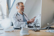 © Mareen Fischinger/Westend61 - Mature doctor with document smiling at video call on computer in doctor's office