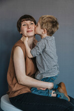 Cute Boy Kissing Mother On Cheek Over Gray Background
