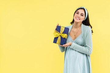 cheerful pregnant woman smiling at camera while holding gift isolated on yellow.