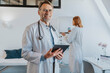 © Mareen Fischinger/Westend61 - Smiling doctor with digital tablet standing with coworker in background at clinic