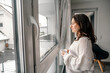 © Oxana Guryanova/Westend61 - Woman standing with coffee cup looking through window at home