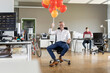 © peter scholl/Westend61 - Smiling businessman holding bunch of balloons while sitting on chair in office