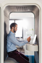 Male Entrepreneur Attending Meeting On Laptop In Telephone Booth At Office