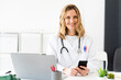 © Giorgio Fochesato/Westend61 - Smiling female medical worker with smart phone and laptop sitting at desk in medical clinic