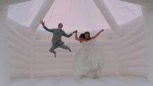 A Playful Bride And Groom Jump In Unison In A Bouncy Castle As They Pose For Some Fun Photos.