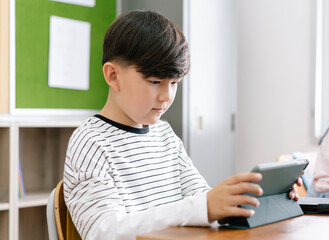 Wall Mural - Portrait of pupil little boy concentrating with digital tablet while sitting at desk with classmates in classroom at elementary School. Education, Technology and Internet concept