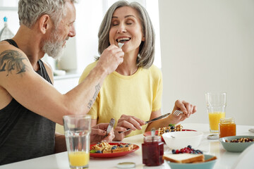 Wall Mural - Older husband feeding mature wife tasting waffles having breakfast at home. Happy healthy affectionate senior couple eating dessert sitting at kitchen table having fun enjoying morning meal together.