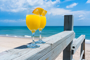 Two glasses with yellow mimosa cocktail. Alcohol drink with bubbles glows in the sun on the background of the beach by the ocean 