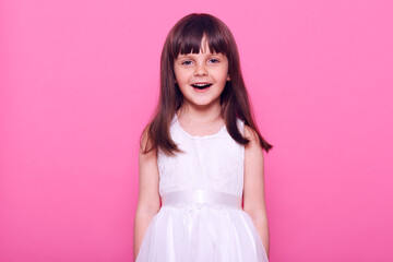 Wall Mural - Cute smiling girl wearing white dress looking directly at camera with happy facial expression, having dark hair, positive mood, isolated over pink background.