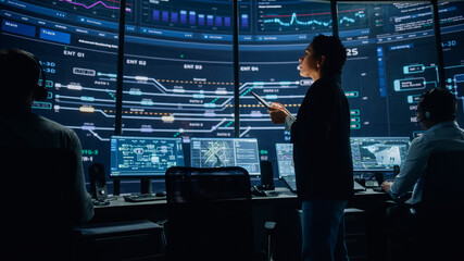 Fototapete - Young Multiethnic Female Government Employee Uses Tablet Computer in System Control Monitoring Center. In the Background Her Coworkers at Their Workspaces with Many Displays Showing Technical Data.